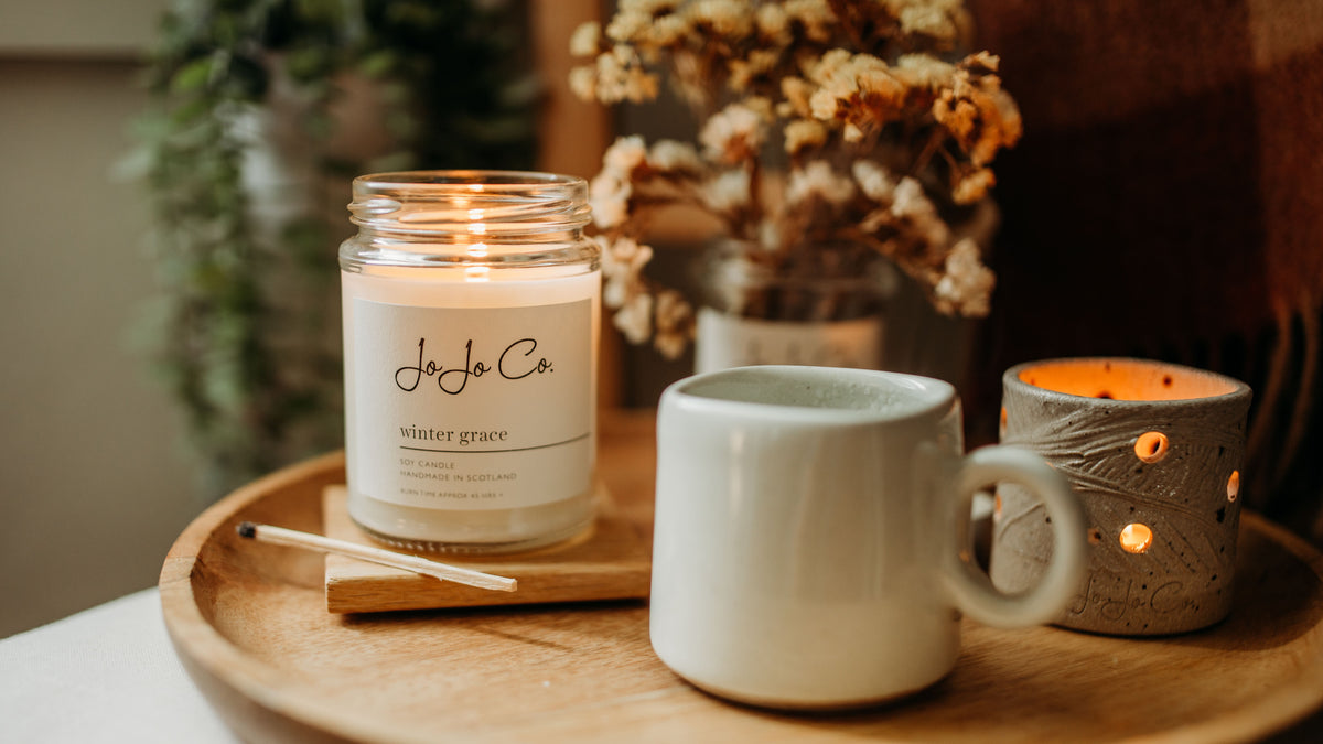 Winter Grace soy candle burning beside a ceramic mug and glowing tealight holder, styled with dried flowers on a wooden tray.
