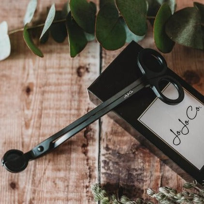Matt black cast iron wick trimmers with a small JoJo Co. logo etched on them sits on top of a black branded box beside a pretty green leaf