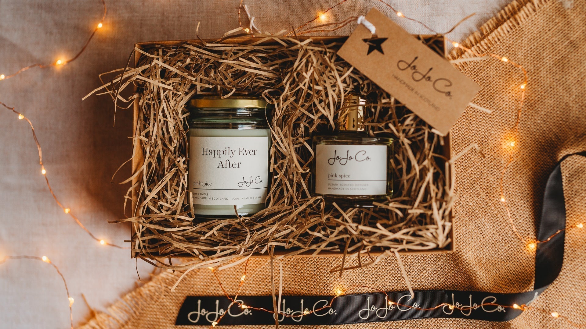 A luxury wedding gift box with a Happily Ever After candle and diffuser, surrounded by warm lights and ribbon in a cosy setting 