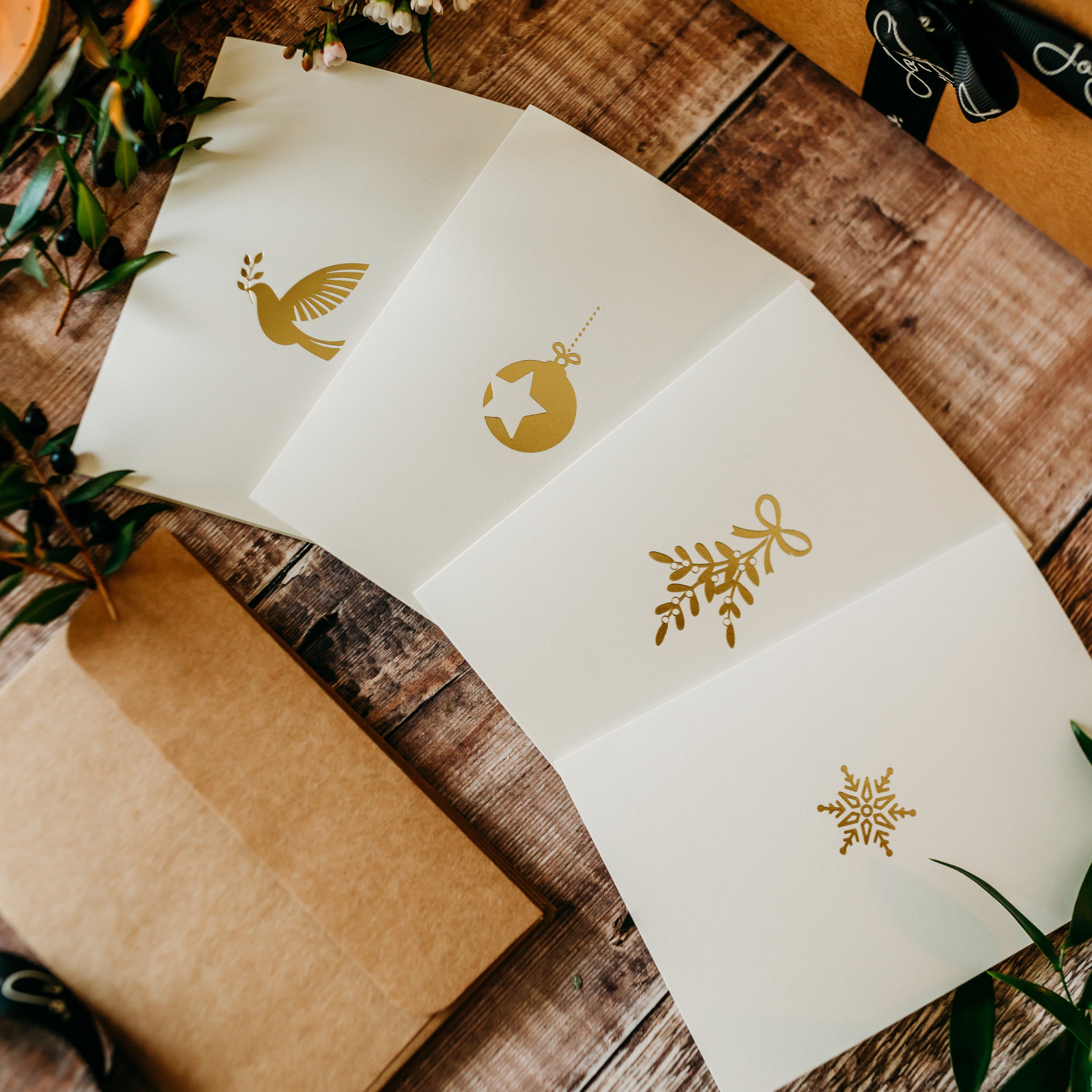 Four cream embossed cards fan out over a wooden table. Each card has a festive themed gold emblem embossed: dove, bauble, mistletoe, snowflake