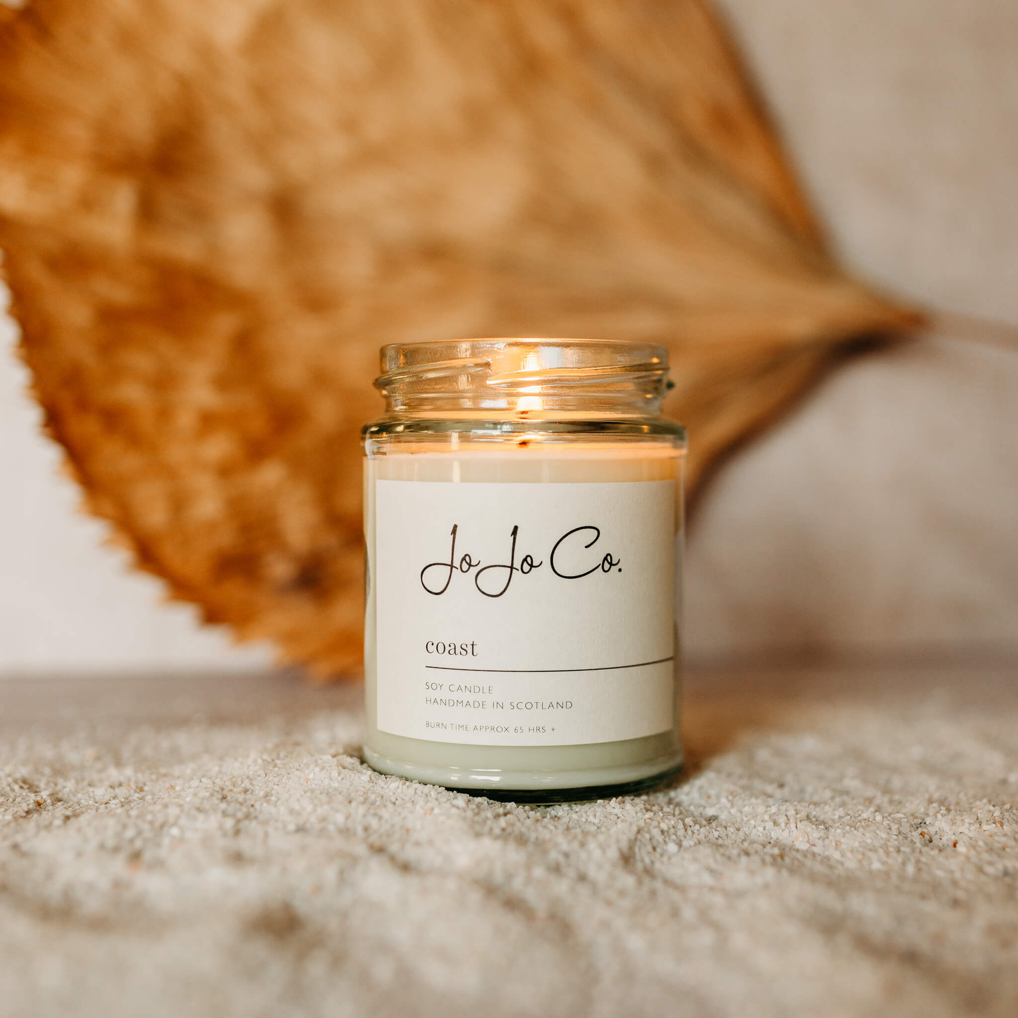 JoJo Co. 65 hour candle with white label and flickering flame. The candle sits on top of sand with a dried leaf in the background.