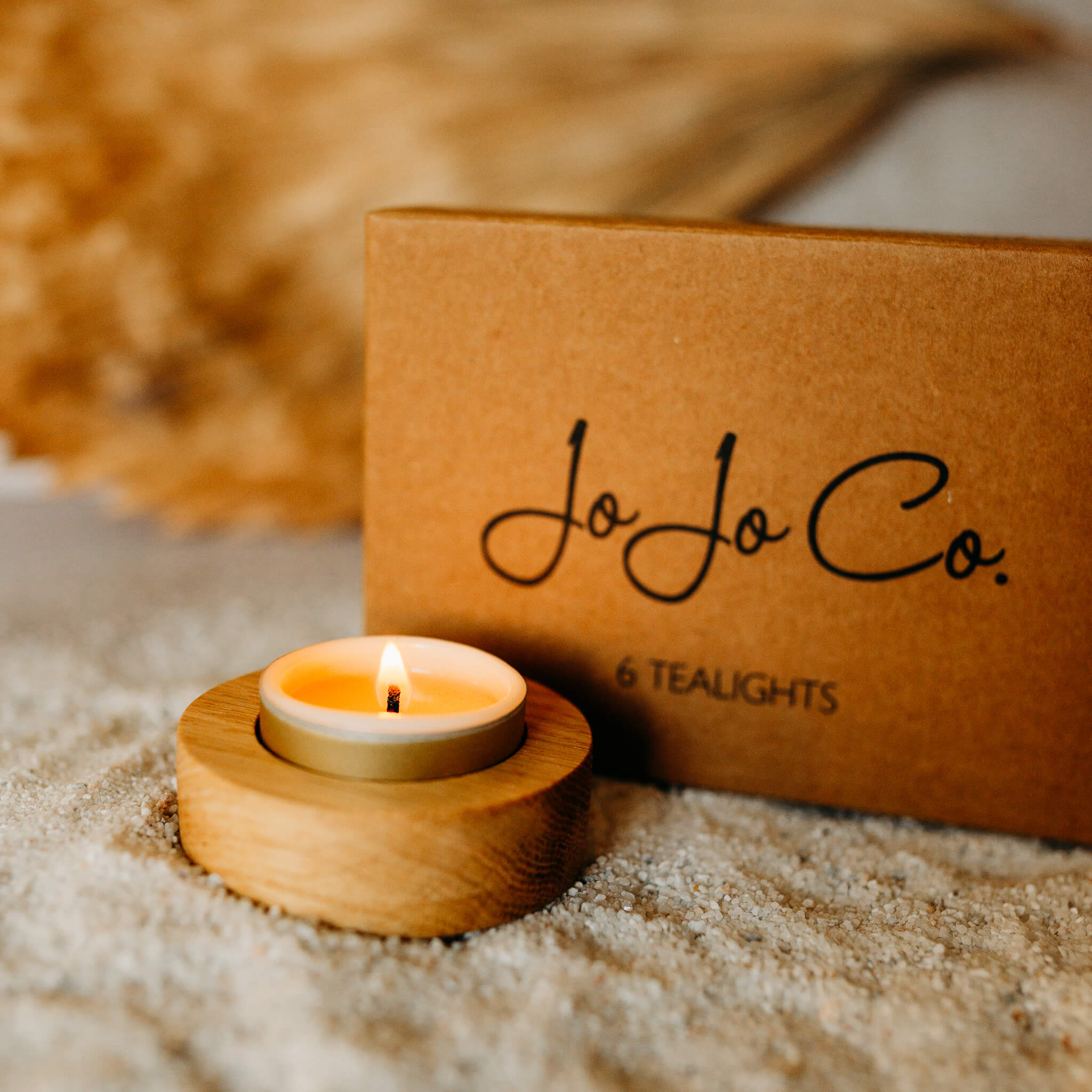 A lit tealight sits neatly in a round oak tealight base, on top of a scattering of sand. In the background is a cardboard box with the words JoJo Co. 6 tealights printed
