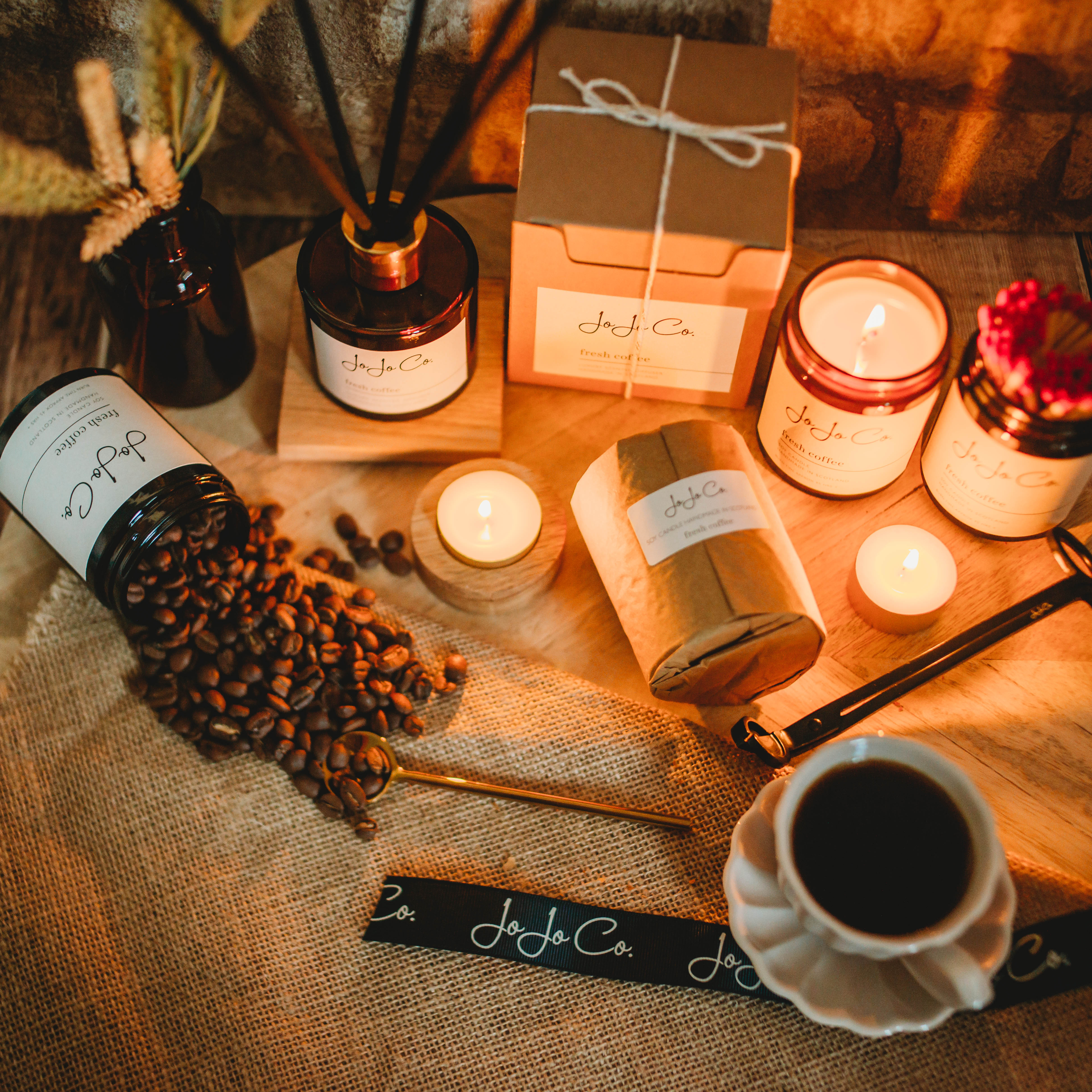 A table with Fresh Coffee diffusers, candles and tealights displayed. Candles are lit, a jar spills coffee beans across the scene and a warm cup of coffee sits on the edge of the scene.