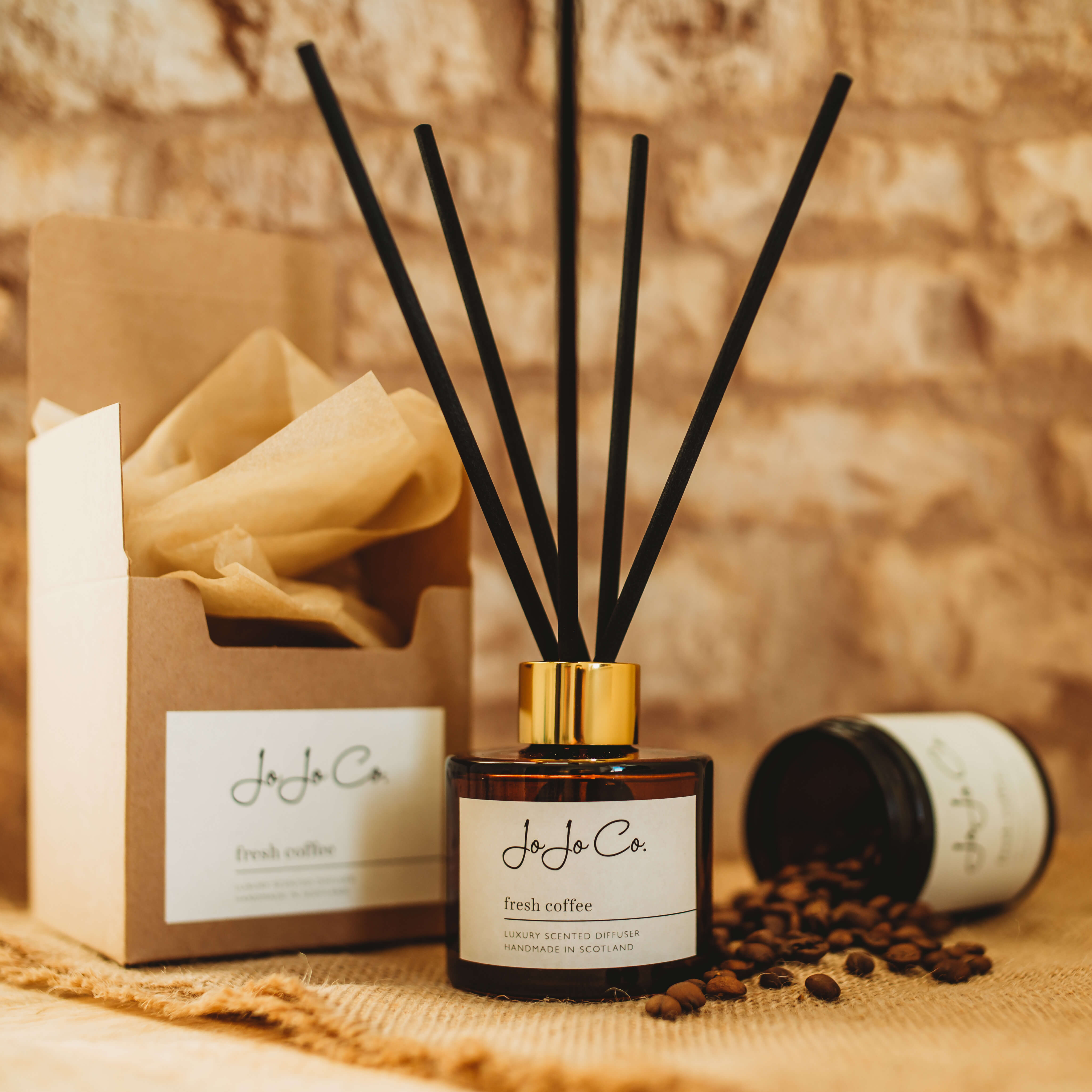 An amber glass jar diffuser with a white JoJo Co. label and gold lid and black reeds sits beside a pretty gift box with tissue paper and a dark jar spilling coffee beans. The label reads JoJo Co. Fresh Coffee