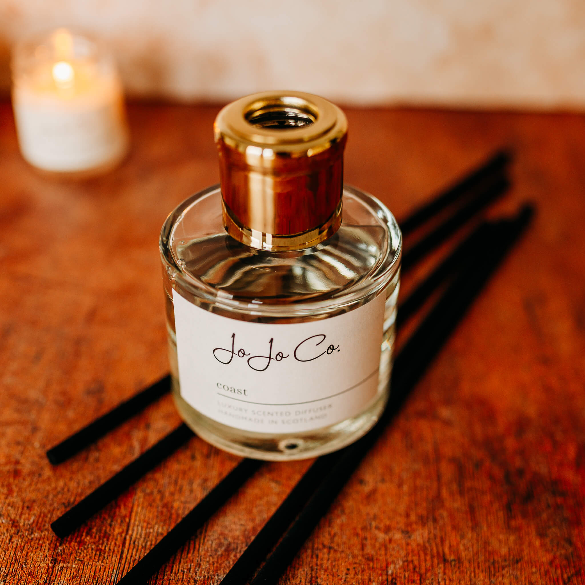 A glass diffuser with white JoJo Co. label and gold lid sits on top of black reeds on a wooden floor. In the background is a blurred lit candle. 