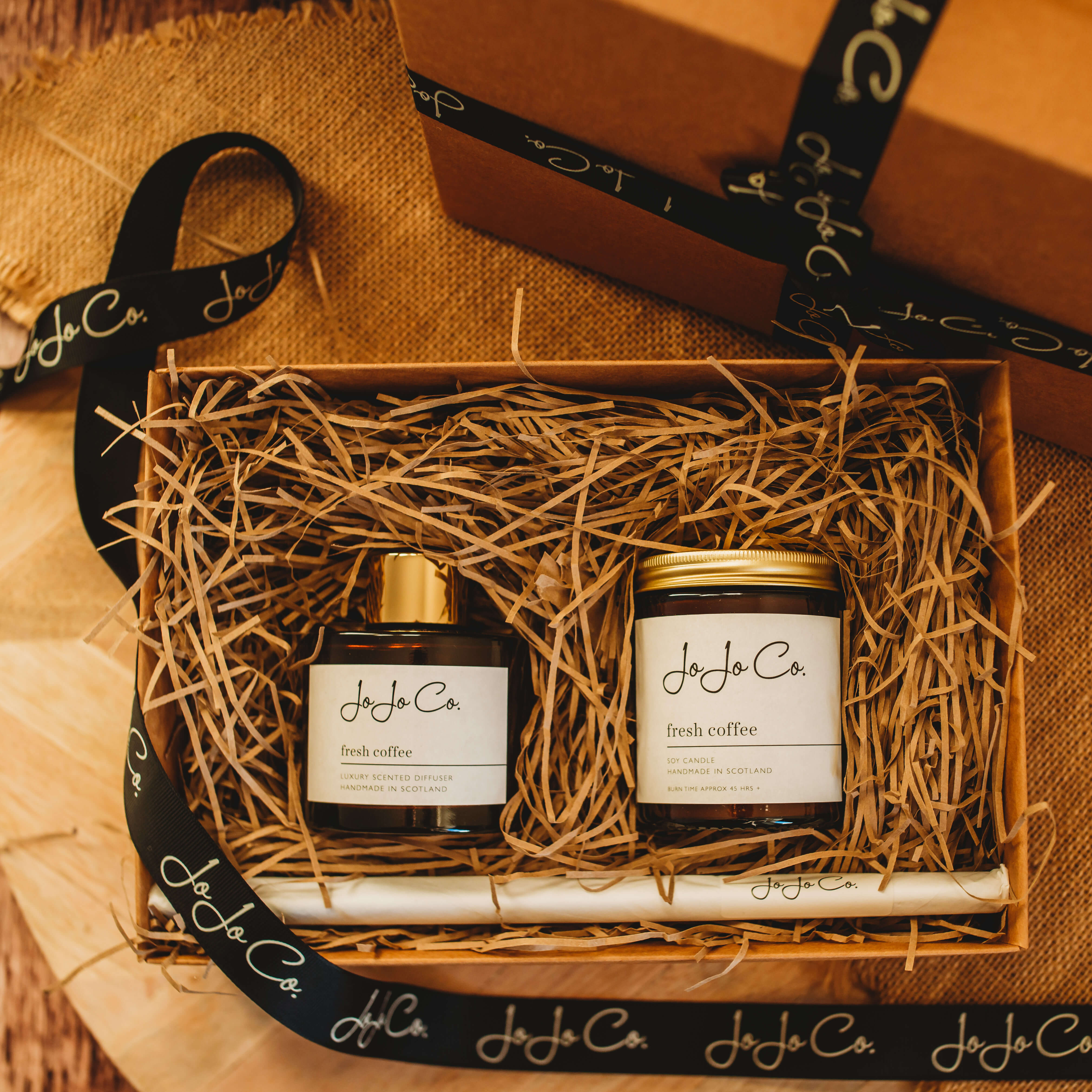 Amber glass jars with gold lids and white JoJo Co. labels are nestled on top of shredded packing paper in a cardboard box. A black tie is draped over it, with a gift box in the background.