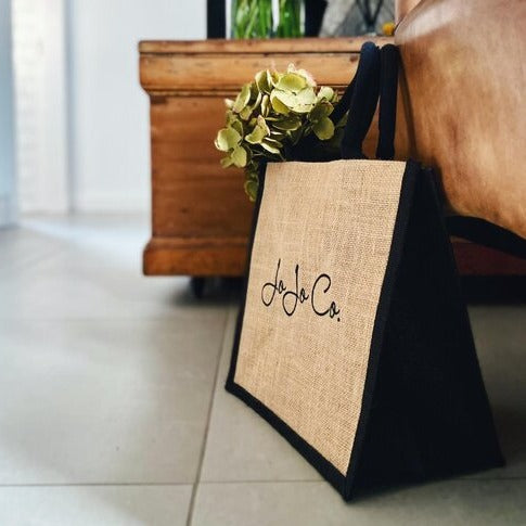 A natural coloured jute bag with black trimming and JoJo Co. logo sits on a tile floor next to brown furniture. A bunch of flowers pokes out of the bag.