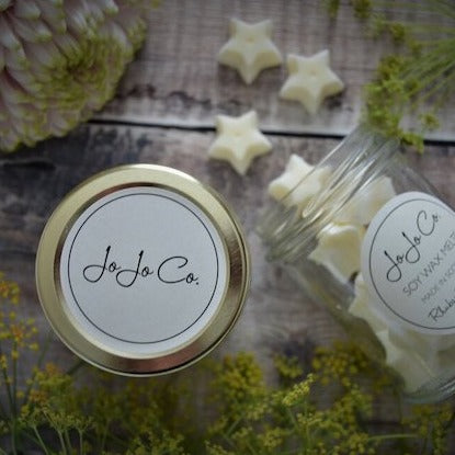 A glass jar with a white JoJo Co. label lies on its side with wax-shaped melts spilling onto a wooden floor. White and green flowers are scattered around, and the lid is at the forefront. 