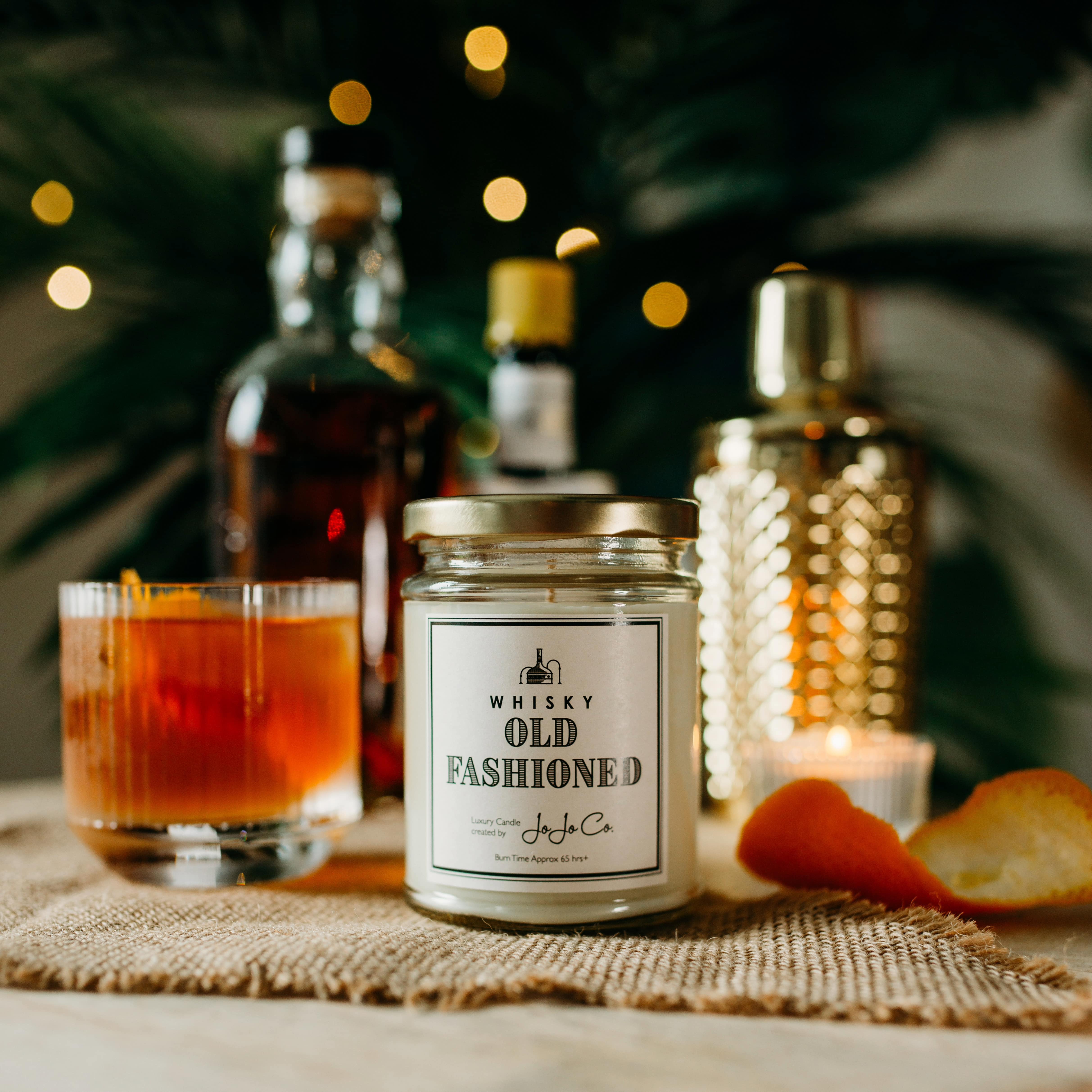 A JoJo Co. Whisky Old Fashioned candle in a glass jar with gold lid and white label sits on a bar. In the background is a large palm, bottles of spirits and a cocktail shaker. Beside the candle is some orange peel and a whisky cocktail.
