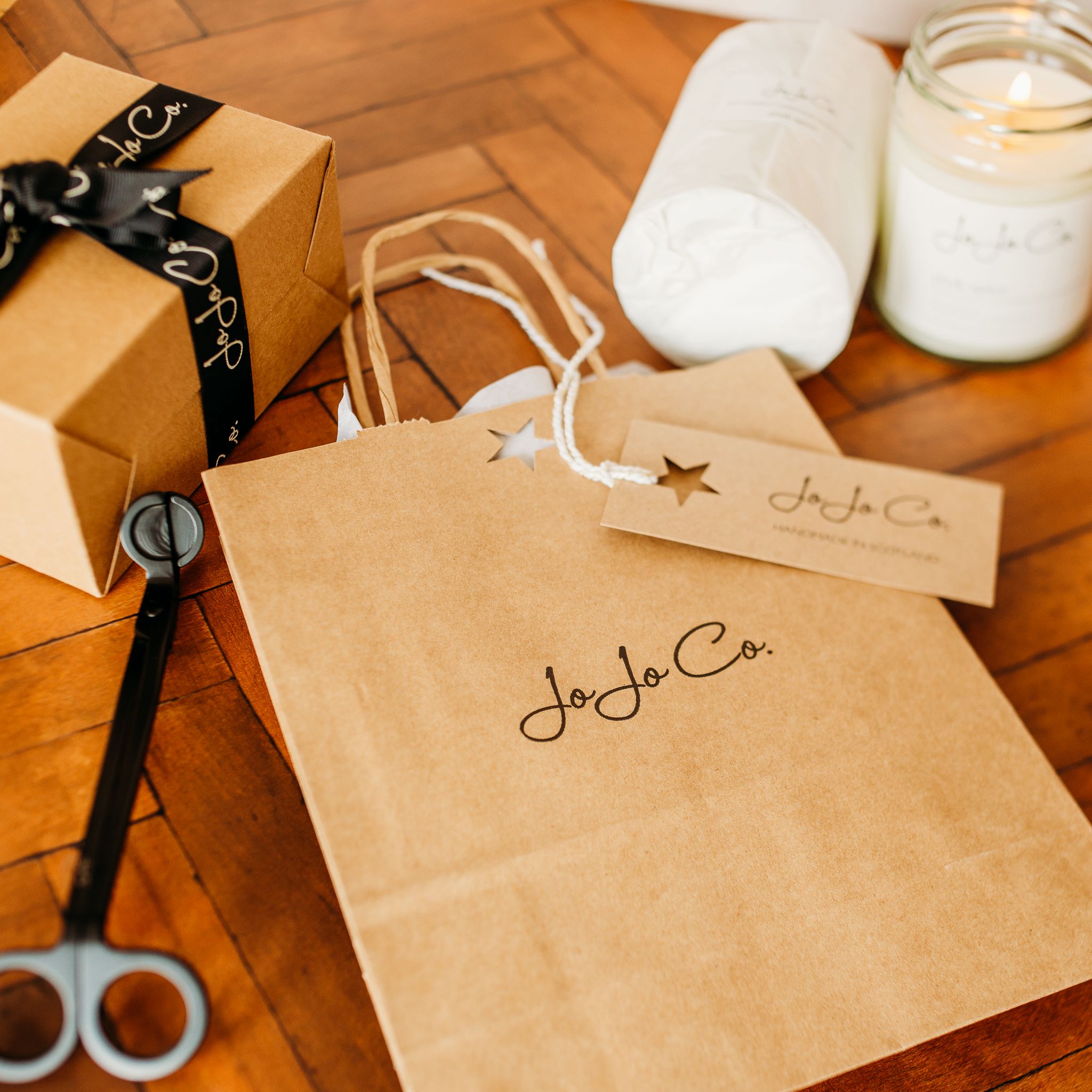 JoJo Co gift bag and tag on a table with candles wrapped in tissue paper in the background and a pair of candle wick trimmers at the side. 