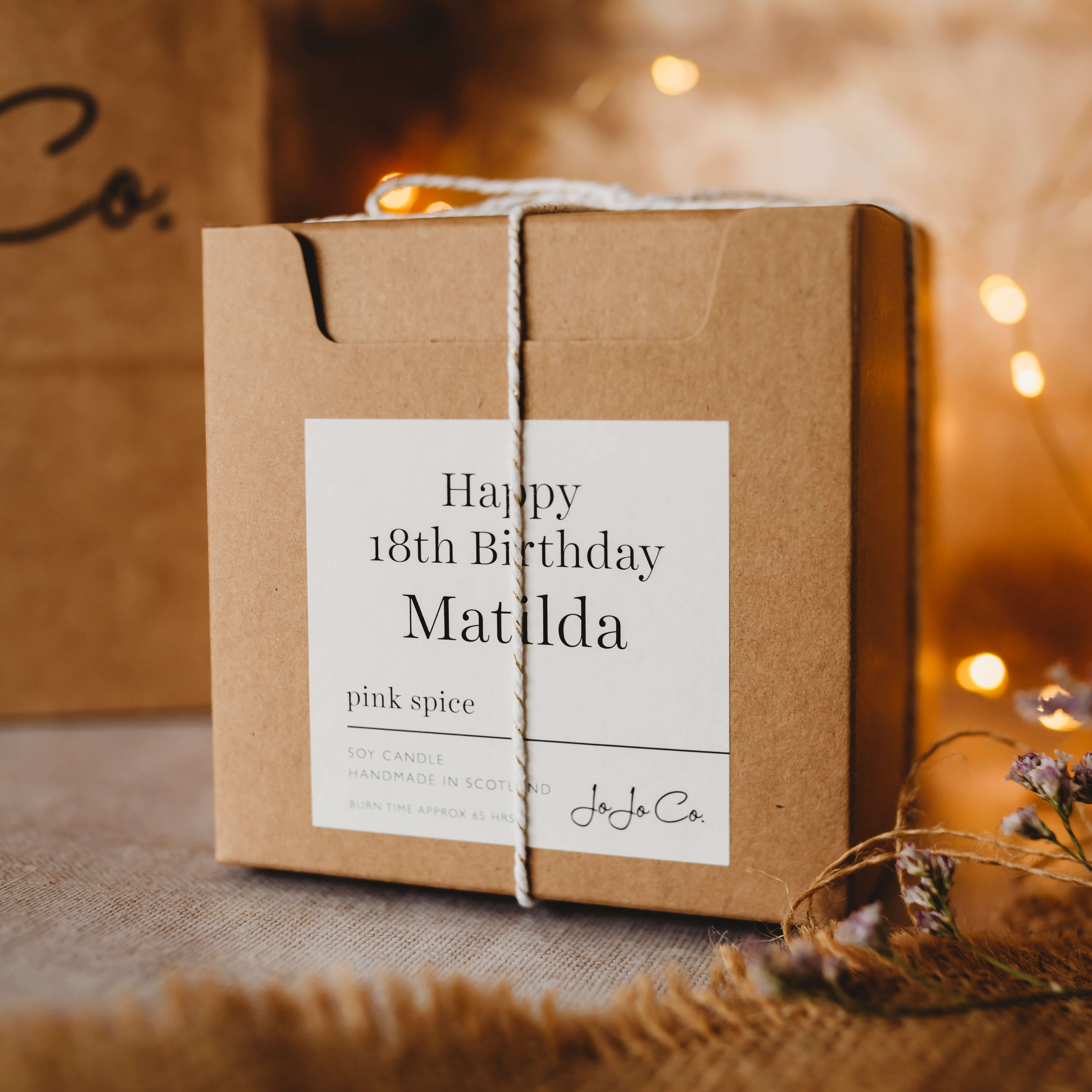 A cardboard gift box with cream label that says 'Happy 18th Birthday Matilda'. Fairy lights are in the background. 