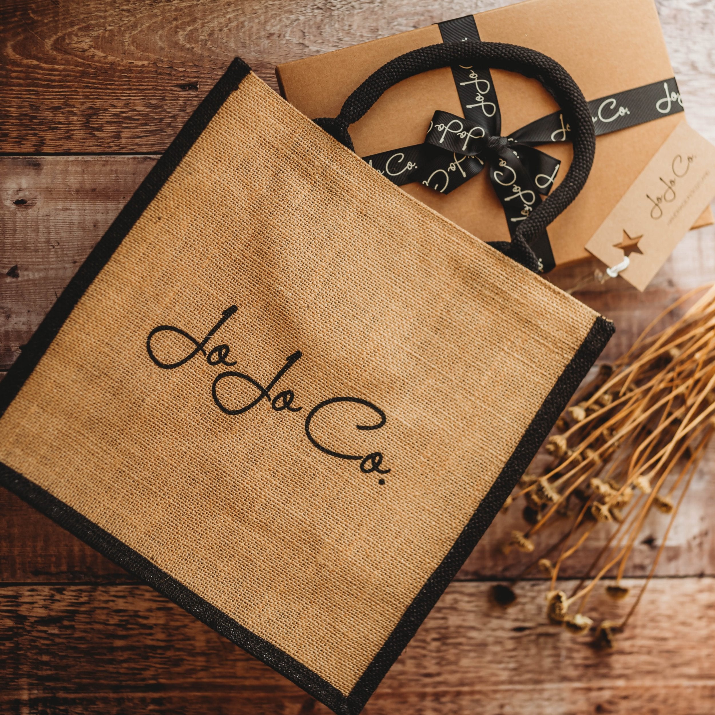 A natural coloured jute bag with black JoJo Co. logo and black trimming sits on top of a cardboard gift box with black branded bow. 