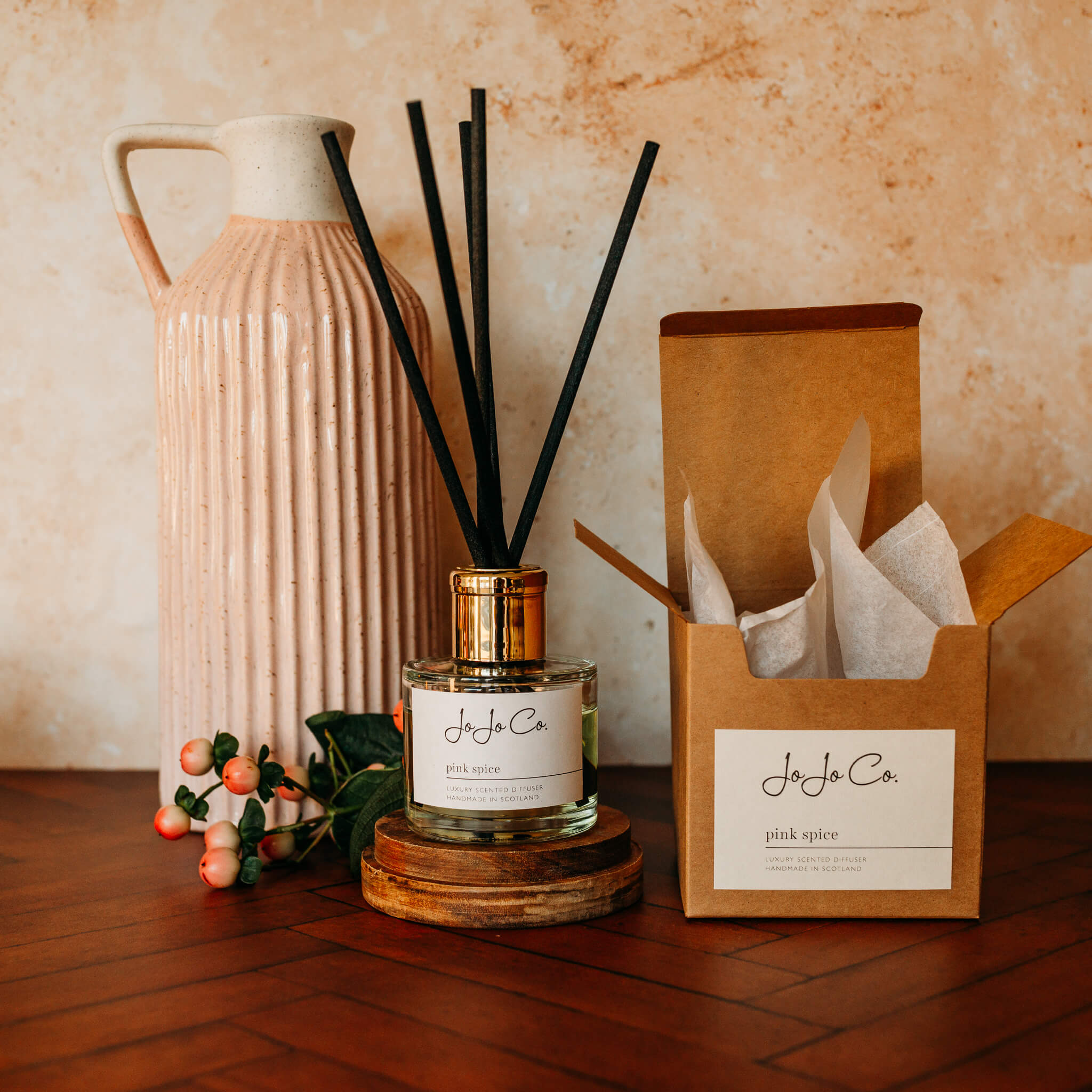 A glass jar diffuser with cream JoJo Co label, gold lid and black reeds sit on an oak candle base on a wooden floor. In the background is a pink and cream jug and pink blush berries on green stalks. 