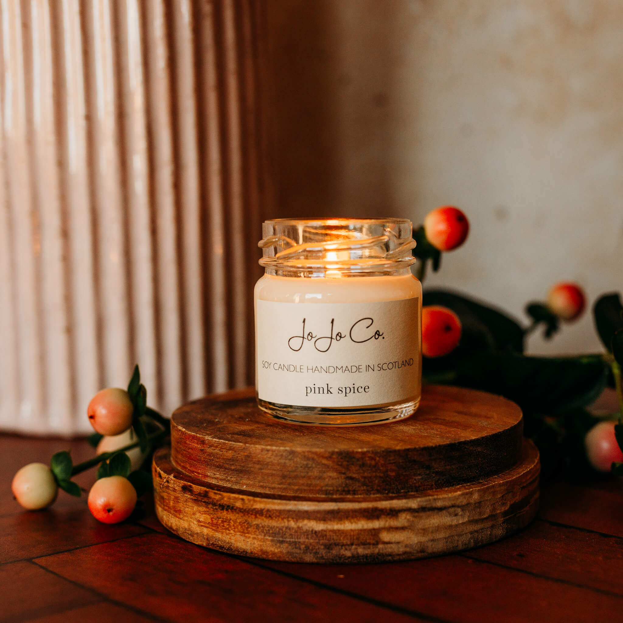 Small JoJo Co. Pink Spice candle sits on a wooden base with pink berries in the background. 