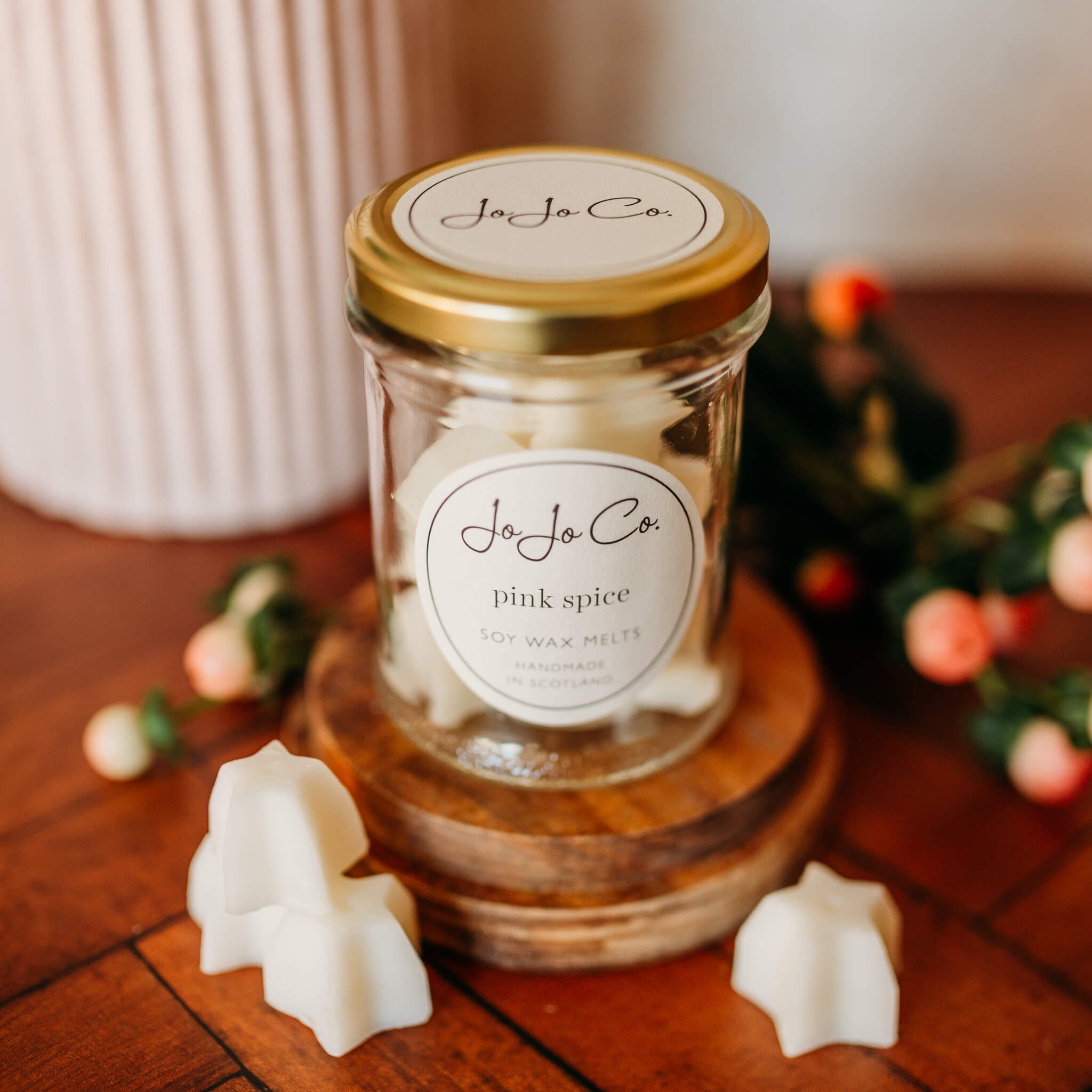 A glass jar with gold lid and white JoJo Co. label sits on a wooden base. White wax melt stars fill the jar and sit beside the jar. In the background are pink berries.