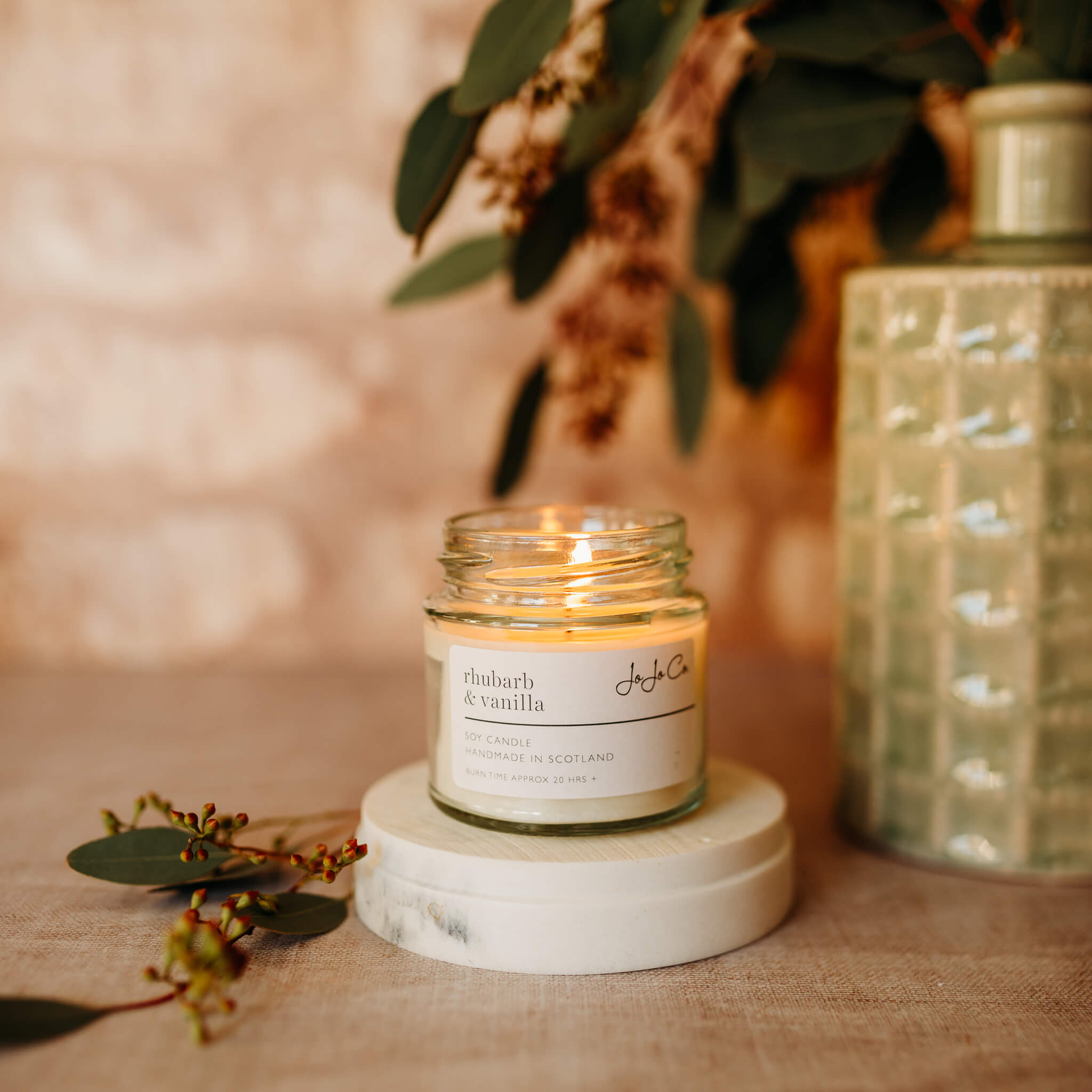 A small, lit candle jar with cream label sits on top of a cream base. In the background is foliage and soft lighting. 