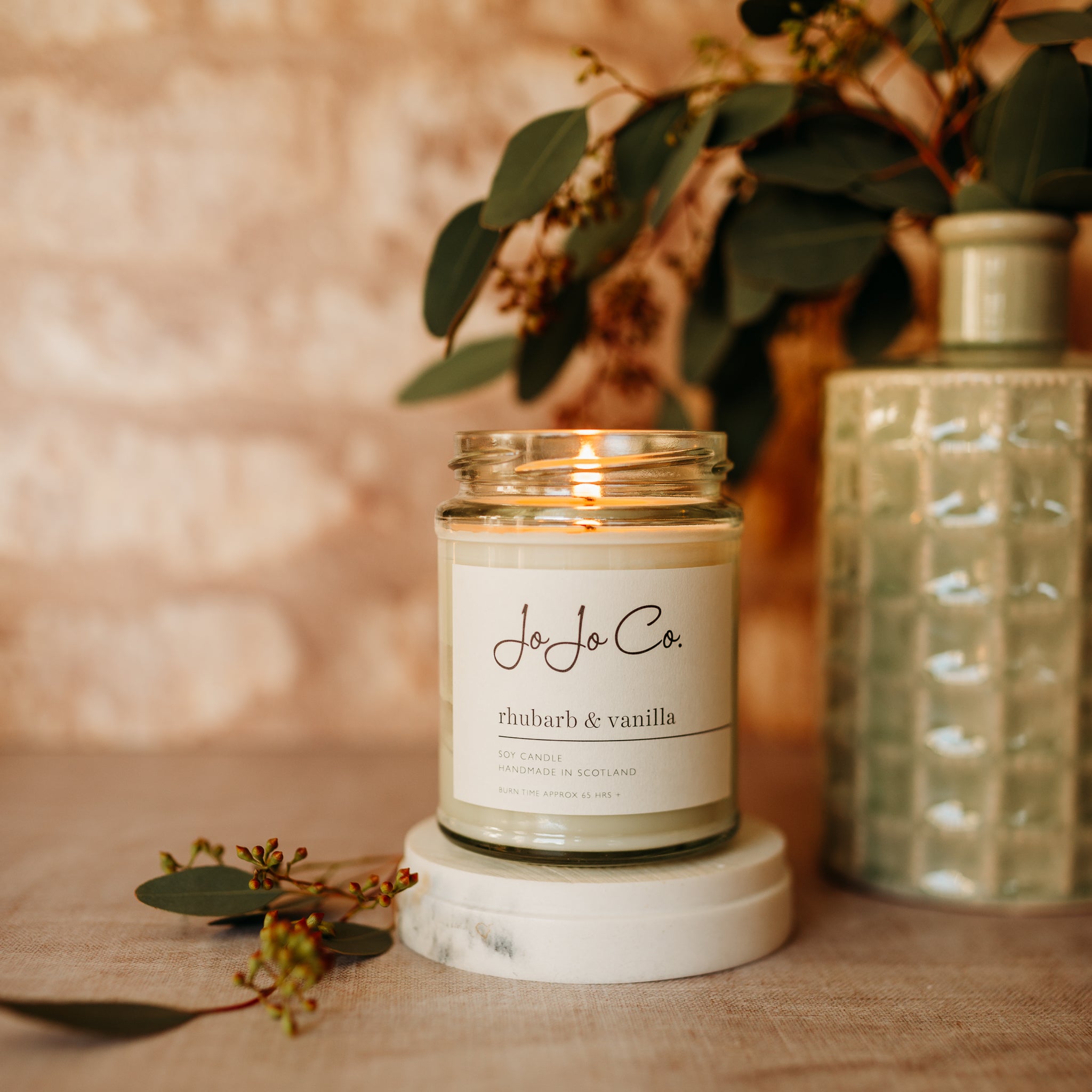 A lit JoJo Co 65 hour Rhubarb and Vanilla candle in a glass jar with a vase and foliage