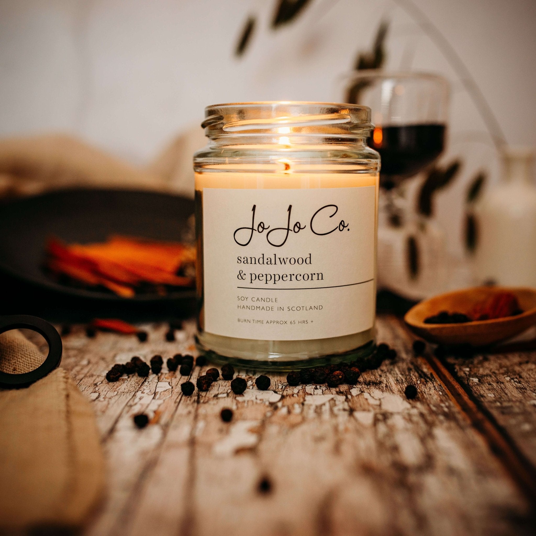 A lit 65 hour Sandalwood and Peppercorn candle in a glass jar with peppercorns scattered in the foreground
