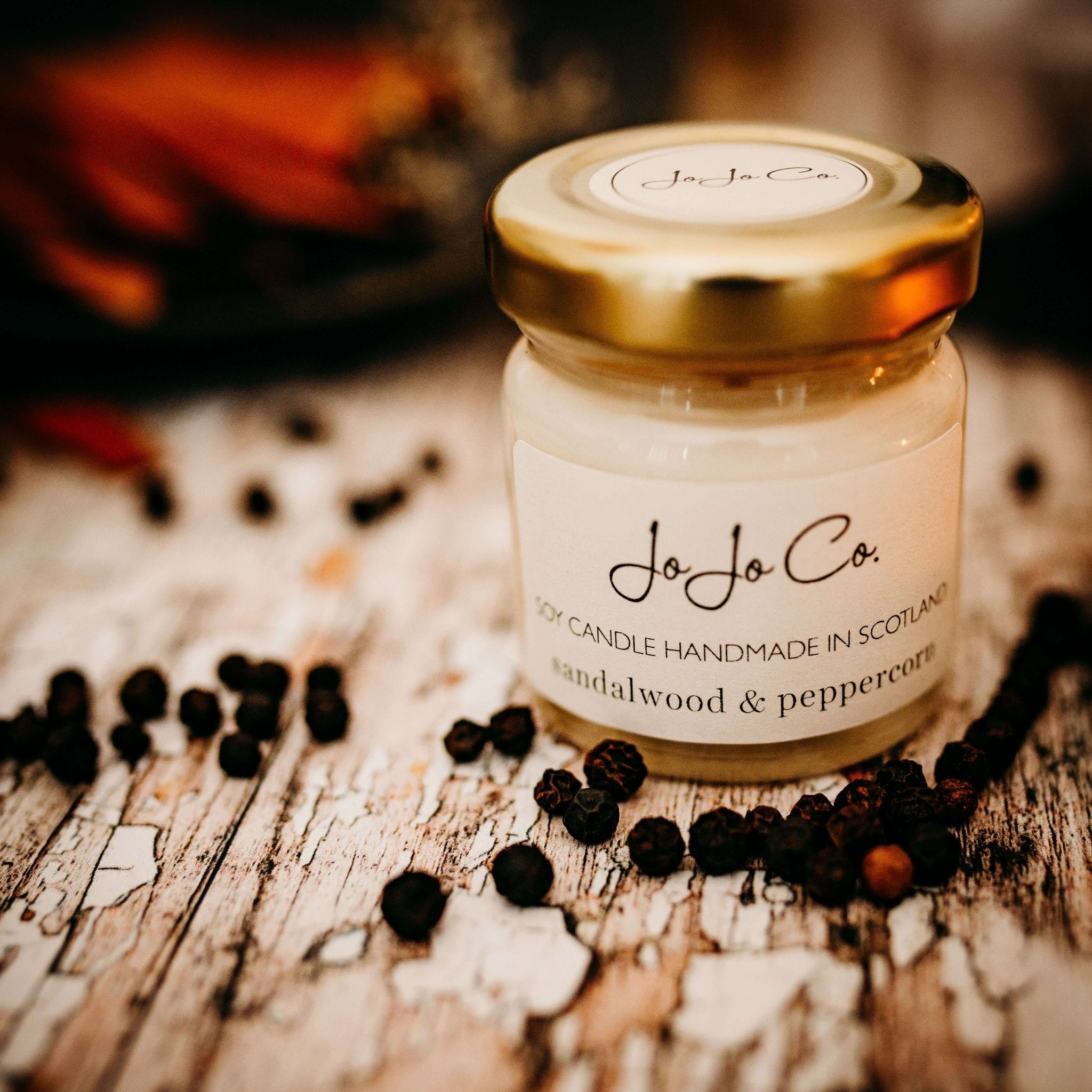 A Sandalwood and Peppercorn mini candle in a glass jar with a gold lid and peppercorns scattered in the foreground.
