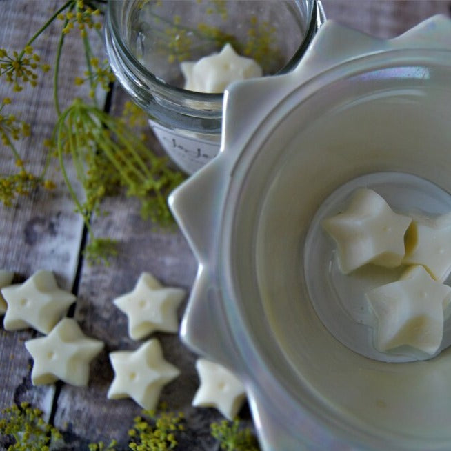 Three white star-shaped melts sit in a pearlescent oil burner. White star-shaped melts are scattered below on a wooden floor, and a jar of melts sits in the background. 