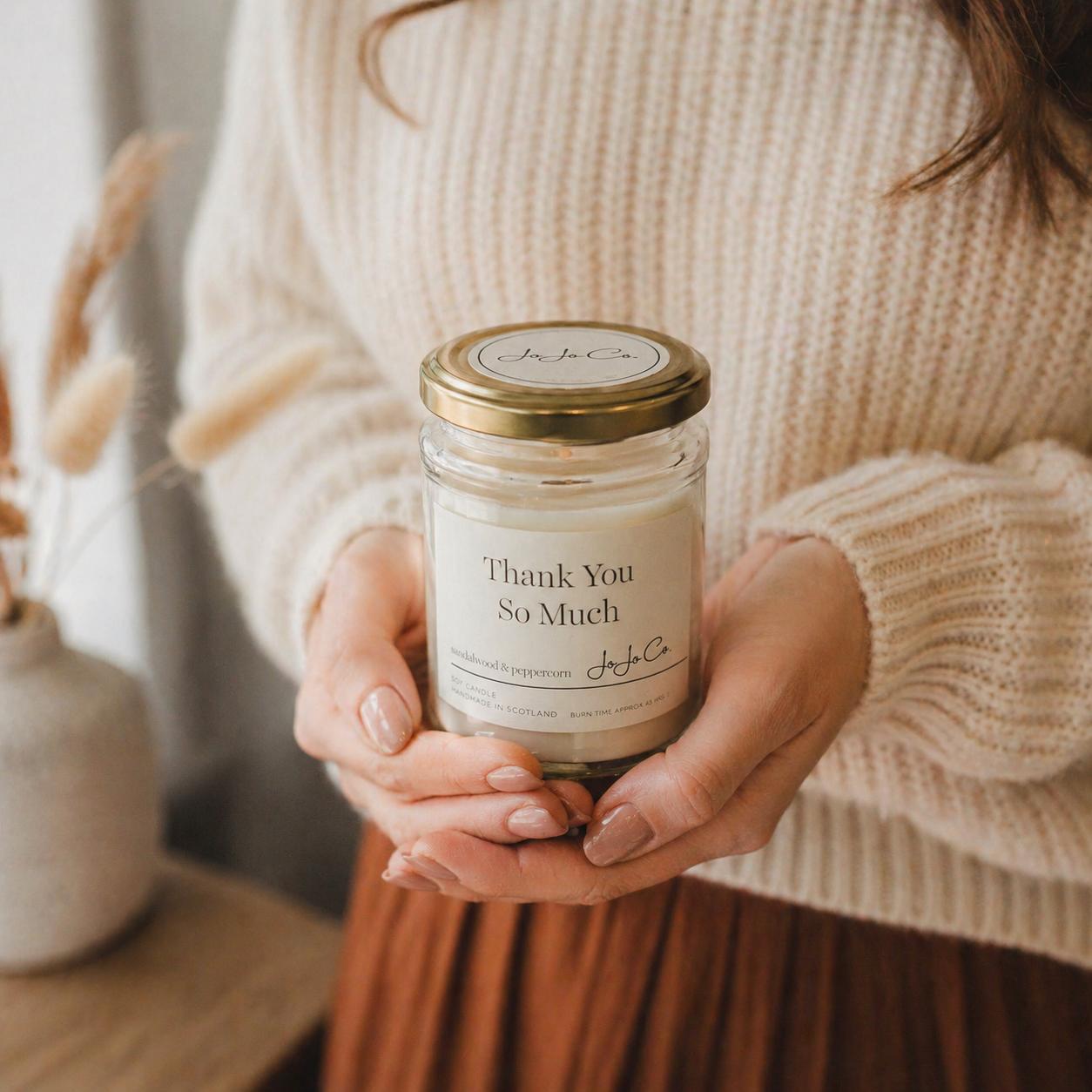 Person holding a candle labeled 'Thank You So Much' in a cozy indoor setting.