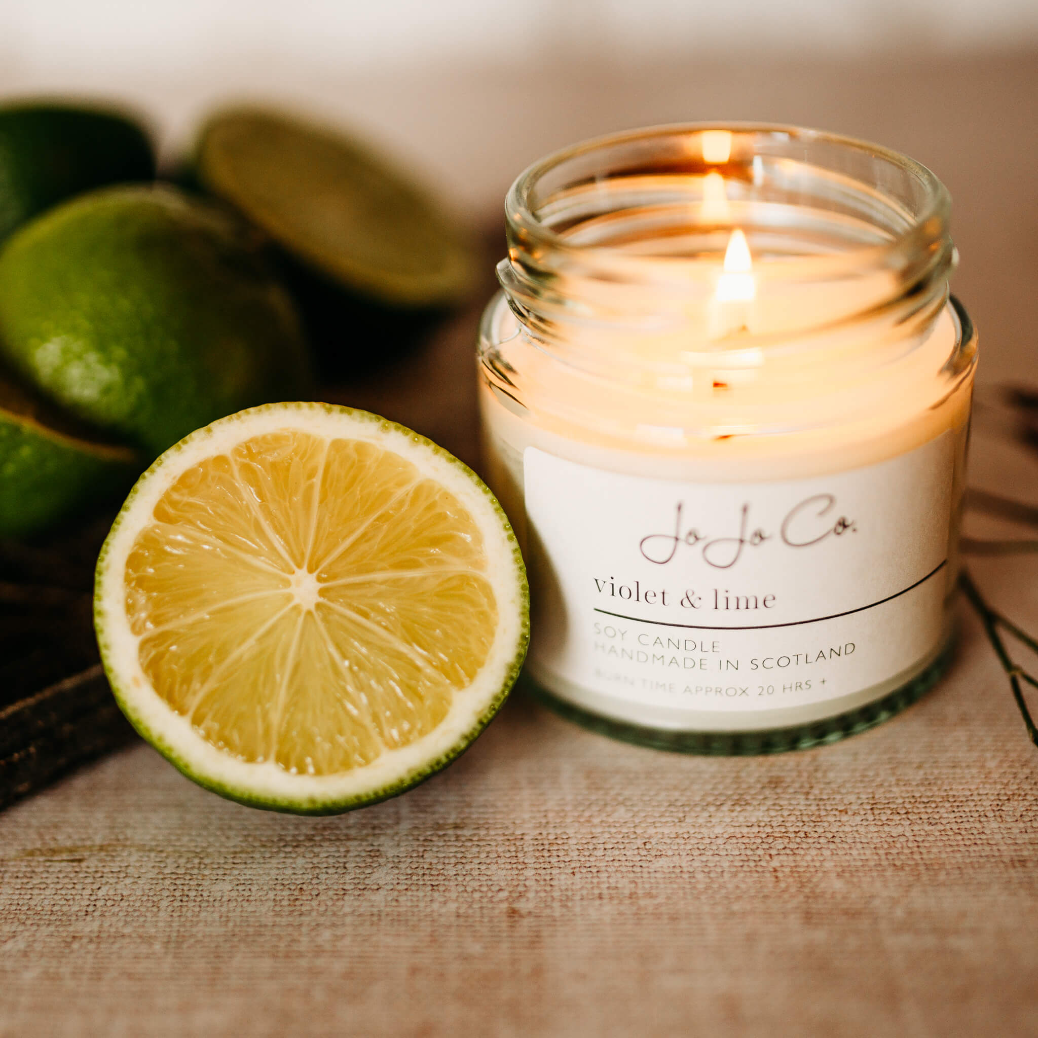 A small JoJo Co. Violet & Lime candle sits beside wedges of lime. The candle is lit, casting a warm glow from the flame.
