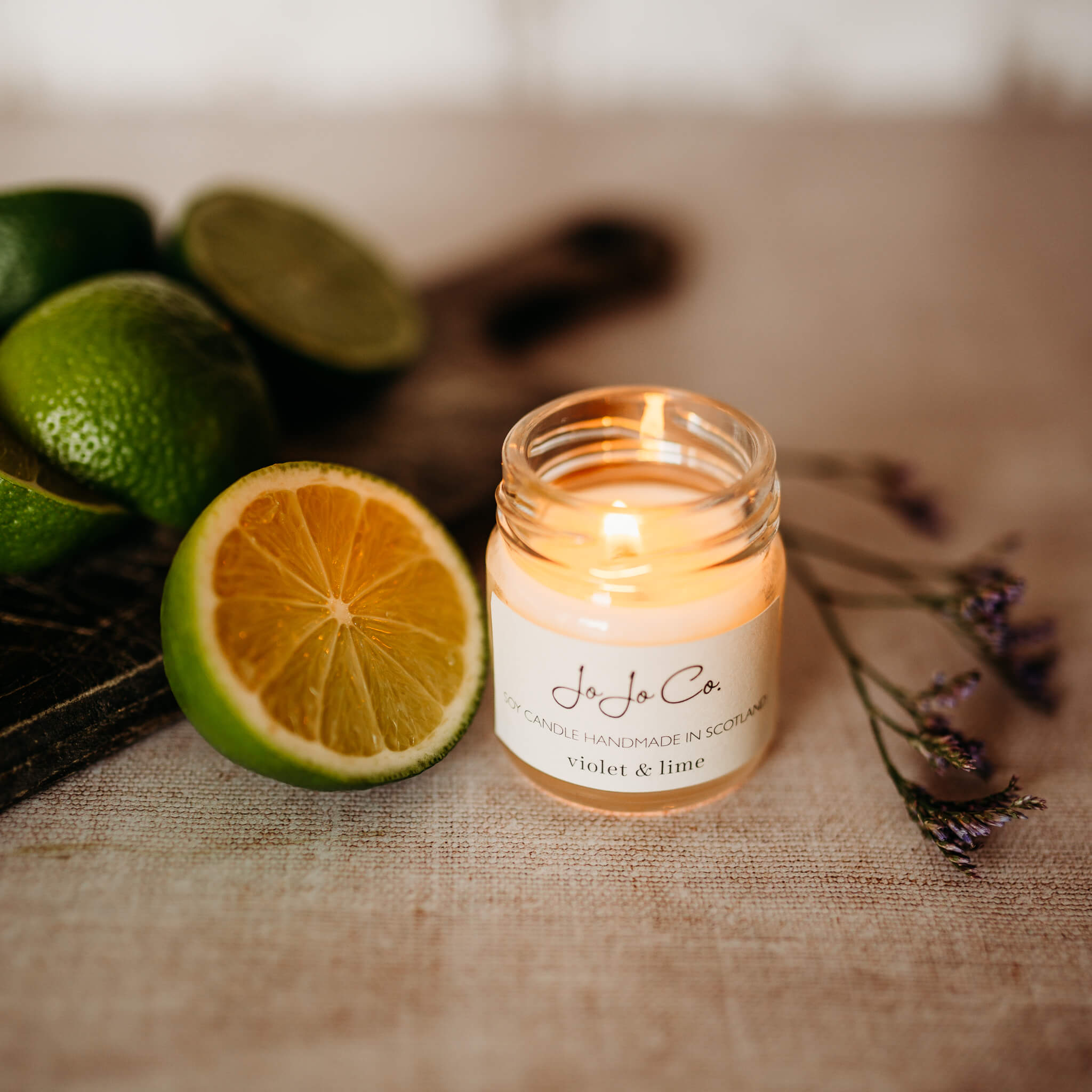 Violet & lime mini candle. A small glass jar from JoJo Co. sits beside slices of lime and dried violets on a jute surface. The candle is lit, casting a warm glow from the glass jar.