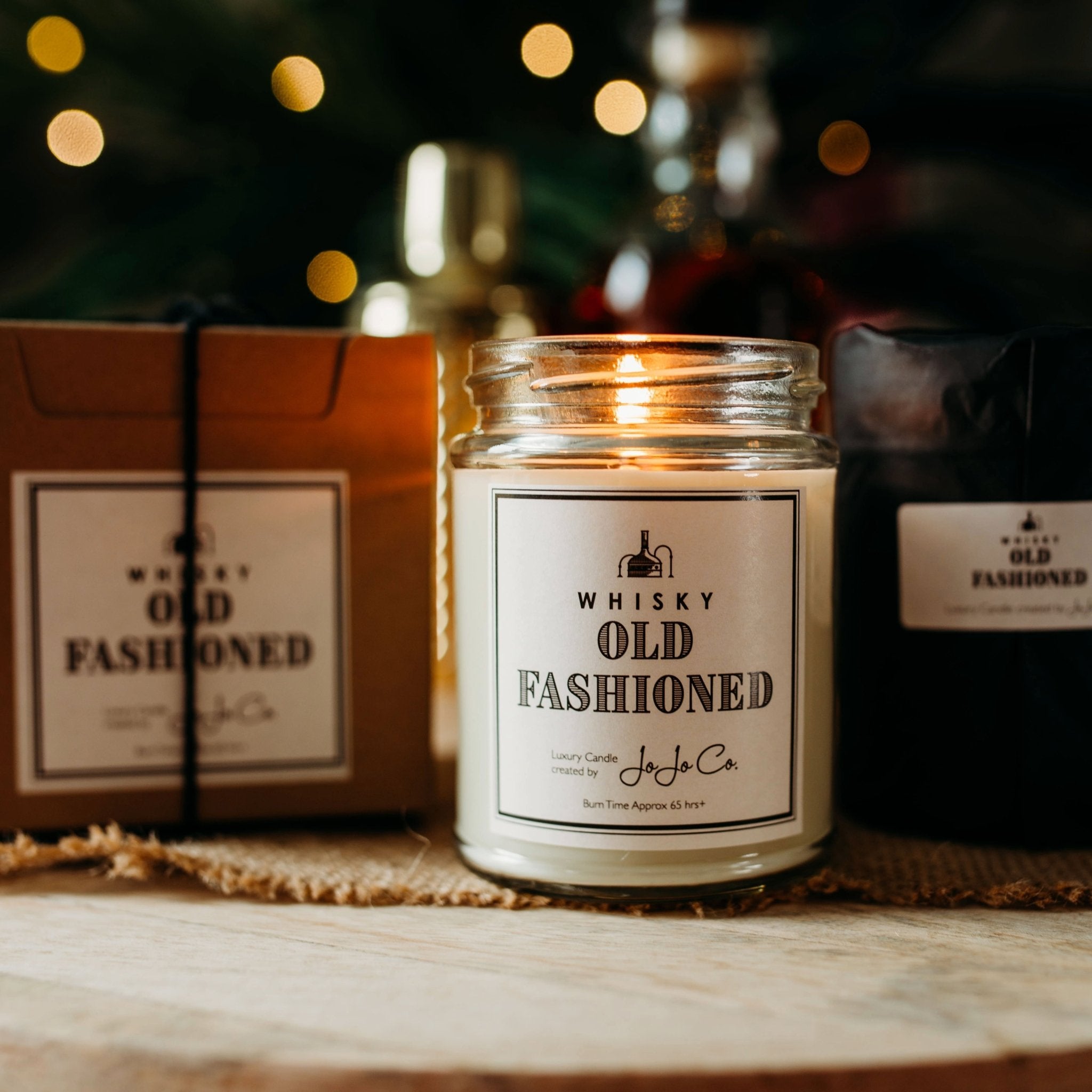 A large JoJo Co. Whisky Old Fashioned candle sits beside a gift box and black tissue paper wrapped candle. Twinkling lights and spirit bottles sit in the background. 