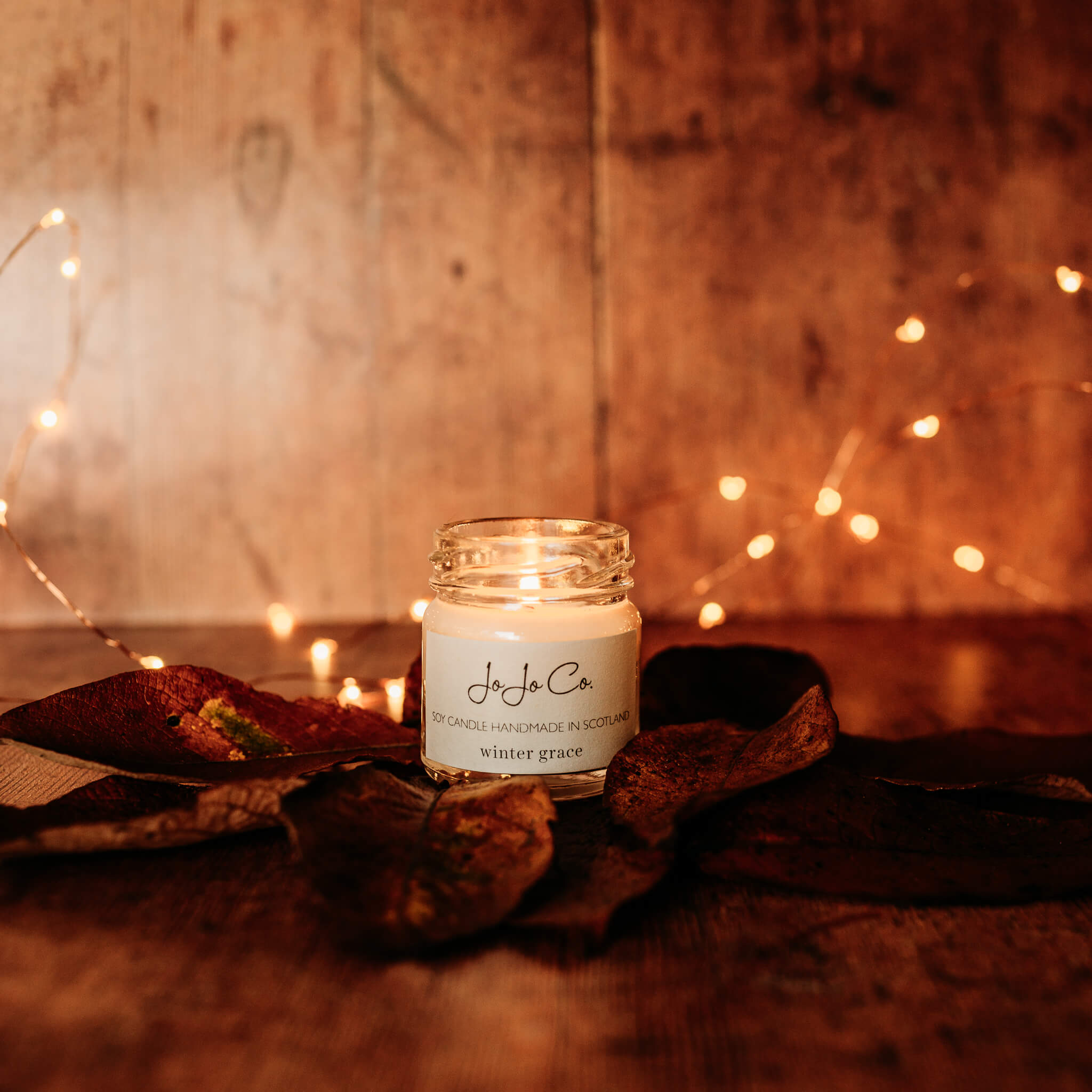 A mini glass JoJo Co. candle with flickering flames. Surrounded by dried leaves and twinkling fairy lights. 