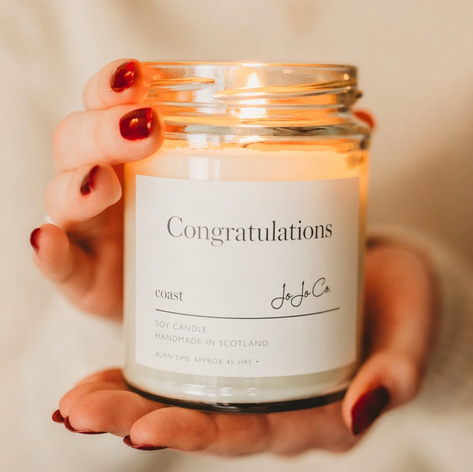 Hand holding a lit candle with 'Congratulations' label against a neutral background
