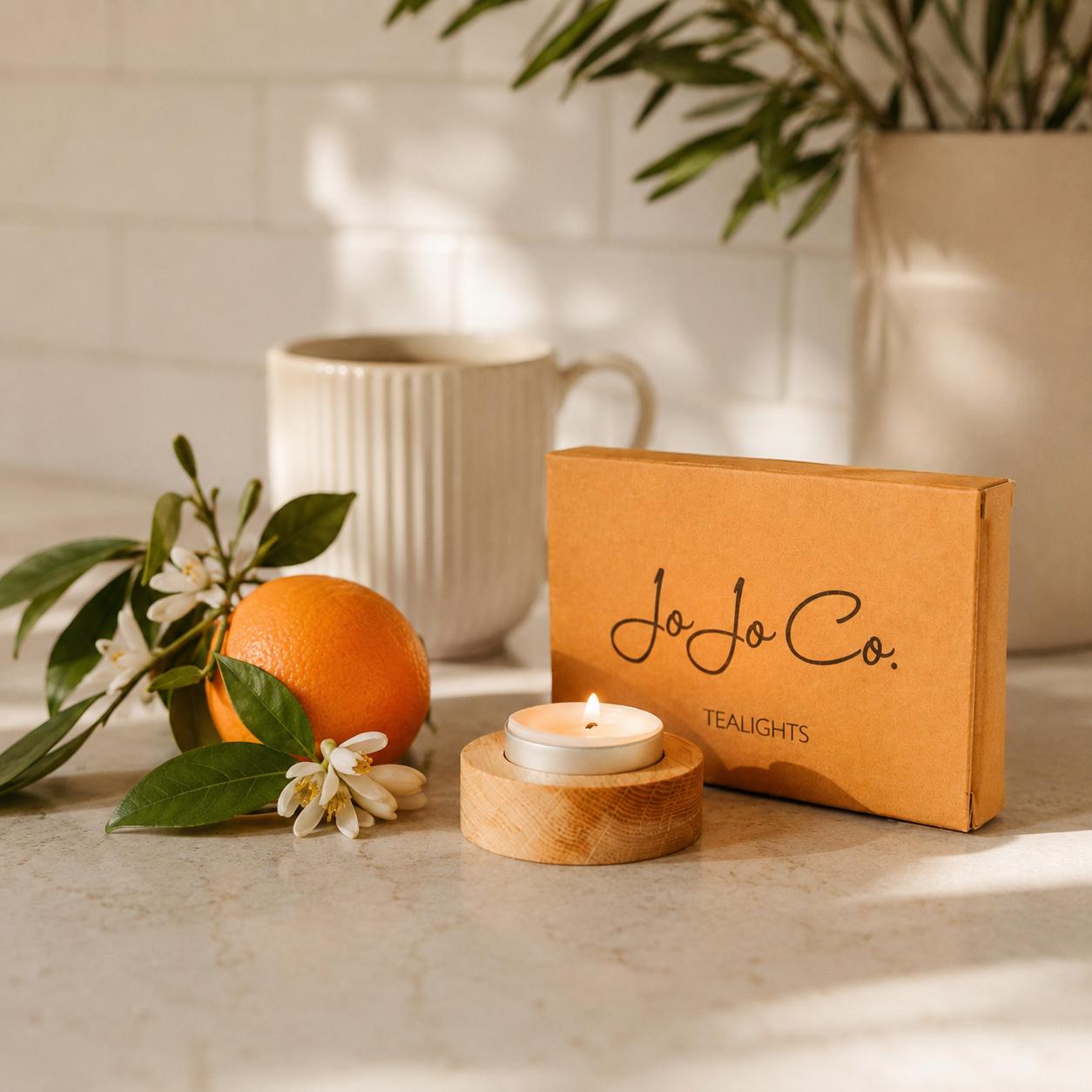 Tealight candle, orange, and flowers on a surface with JoJo Co. packaging in the background.
