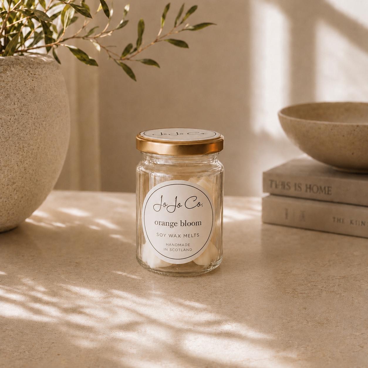 Jar of JoJo Co. orange bloom wax melts on a surface with a plant and books in the background