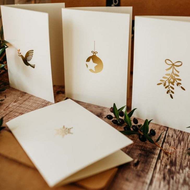 Four cream embossed cards sit on a wooden table. Each card has a festive themed gold emblem embossed: dove, bauble, mistletoe, snowflake