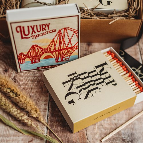Two large boxes of matches sit on a wooden bench. One matchbox is open and red matches are poking out. One box has a picture of the Forth Rail Bridge on it. The other says 'keep the fire burning'
