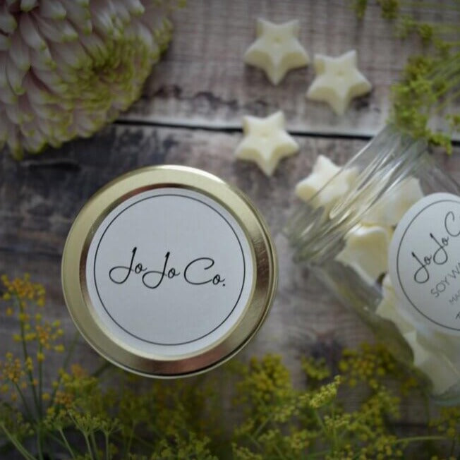Flatlay image of white wax melts spilling from a JoJo Co. jar surrounded by flowers and the lid of the jar with the JoJo Co. label on display